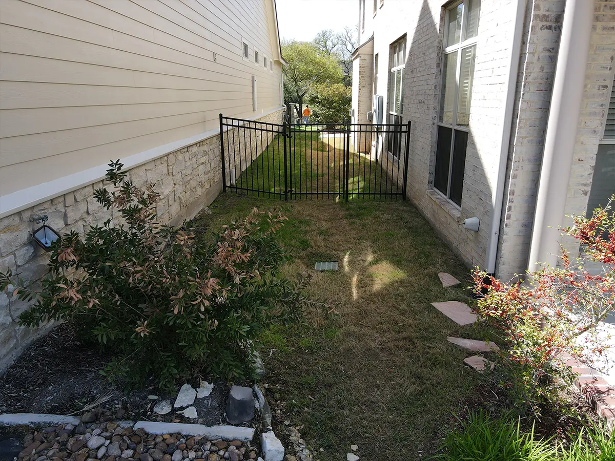 A small aluminum fence between two homes with a dried patch of grass