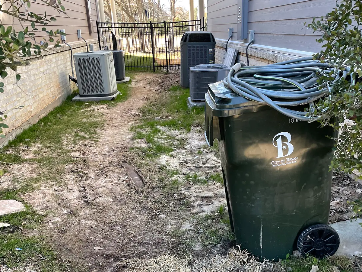 A large trashcan, hose, and HVAC machines between two homes, all placed on mud