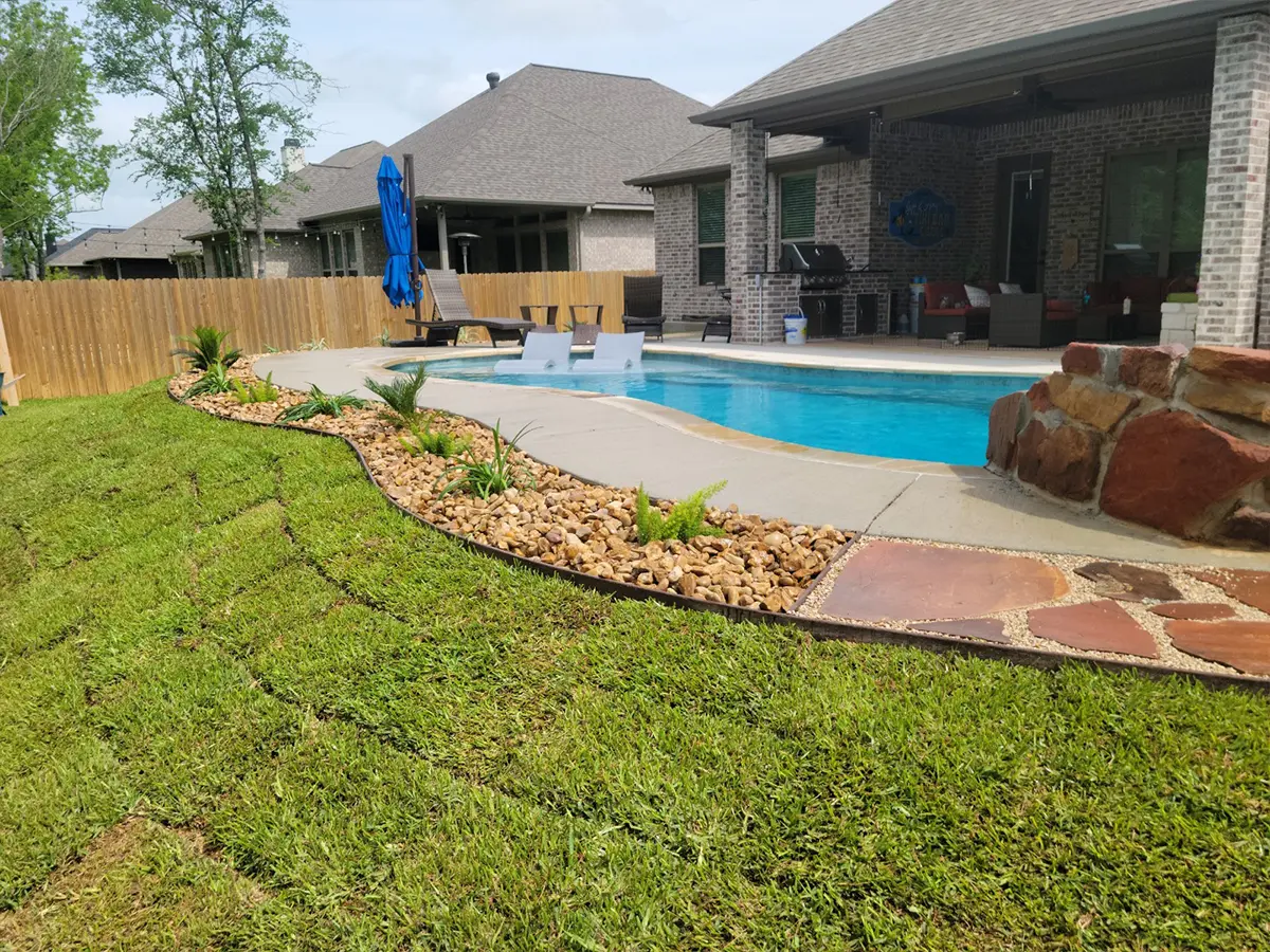 a nice patch of grass around a pool with concrete walkway around it and a small section of plants in pebbles and rocks
