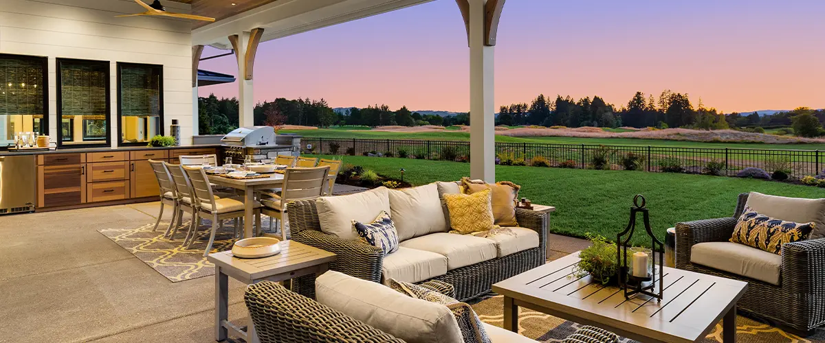 Luxury home exterior at sunset: Outdoor covered patio with kitchen, barbecue, dining table, and seating area, overlooking grass field and trees.