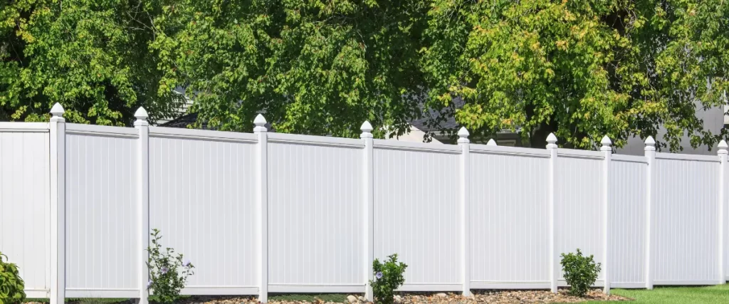 White vinyl fence with lush green trees and small shrubs