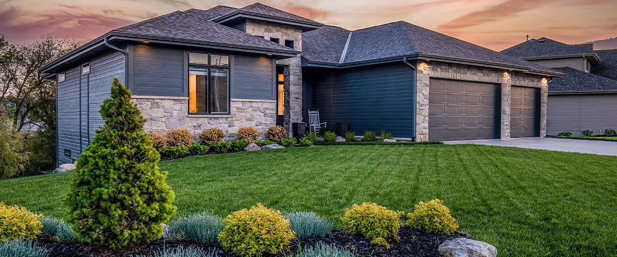 Luxury modern home with stone and dark siding, lush landscaping, and a sunset sky.