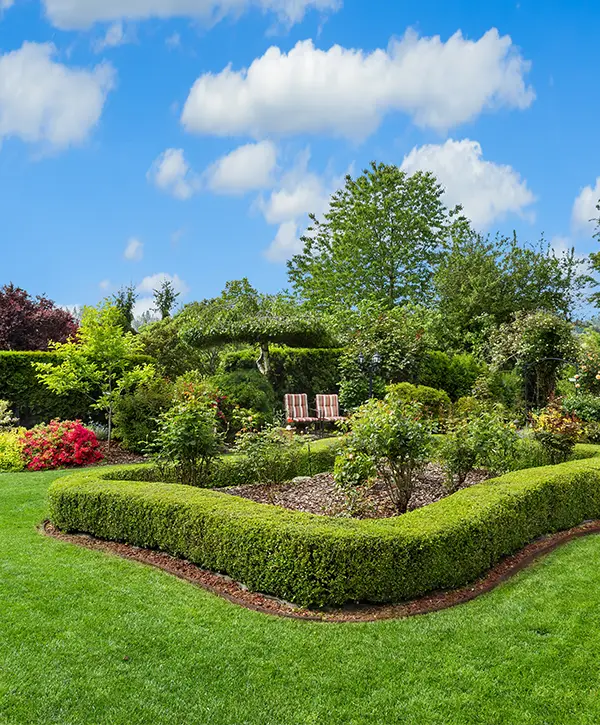 Formal garden landscaping with trimmed hedges, lush lawn, flowering plants, and seating area under a blue sky
