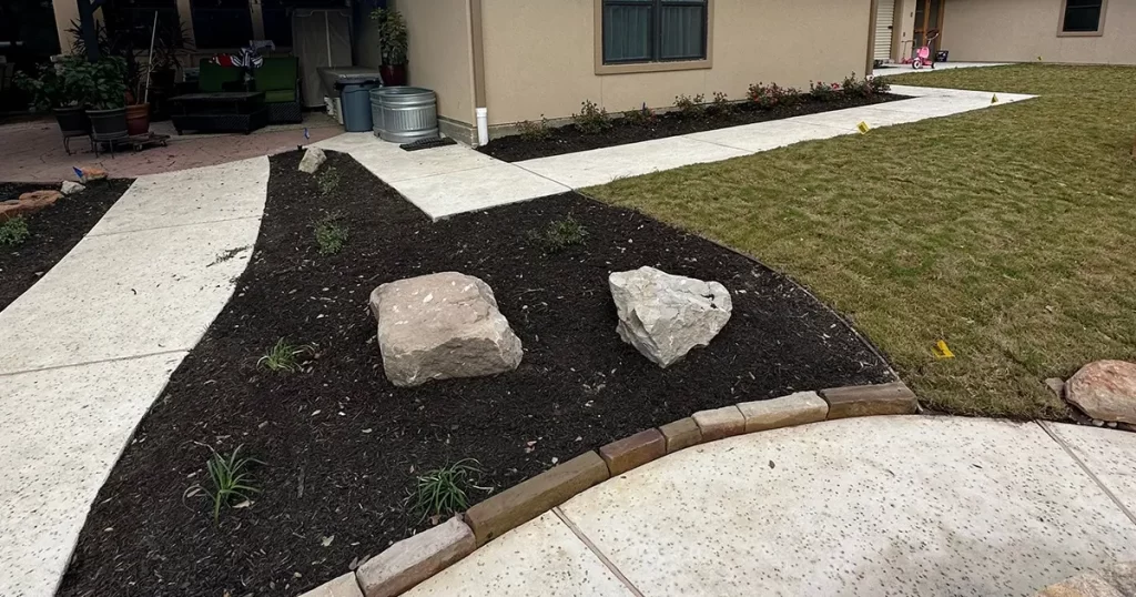 Mulched landscape bed with decorative boulders, concrete walkways, and residential yard landscaping